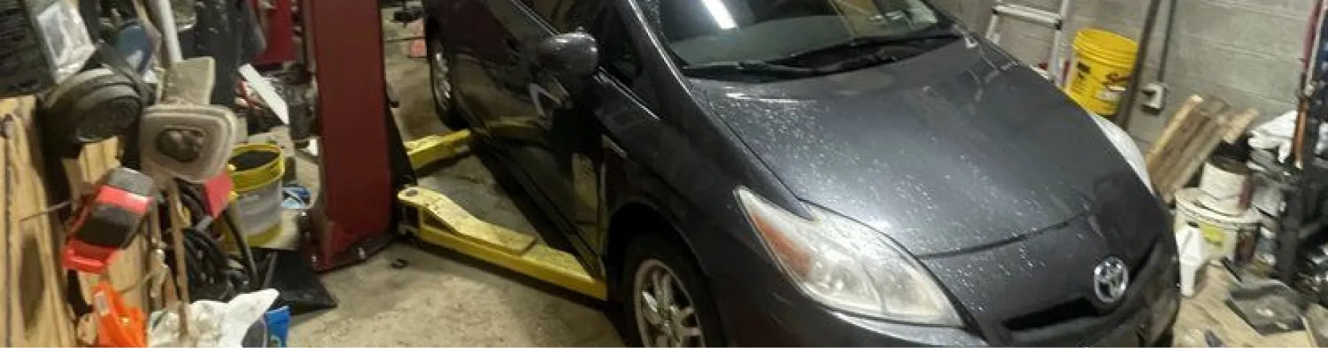 asian auto repair in Canadian Lakes, MI by 2nd Mile Repair. A dark gray Toyota Prius is lifted on a yellow two-post car lift in a cluttered garage.