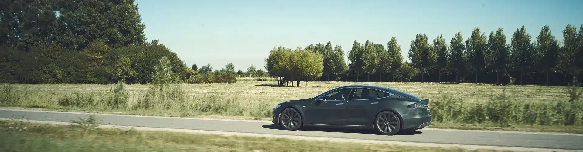 A dark gray Tesla Model S drives on a paved road alongside a grassy field with scattered trees under a clear sky.